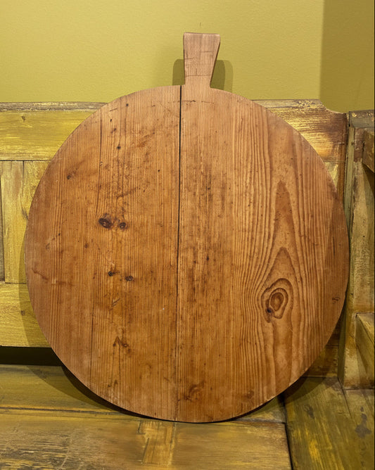A round rustic wooden board with a short handle, leaning against a yellow wall on a wooden bench next to a blue velvet pillow.